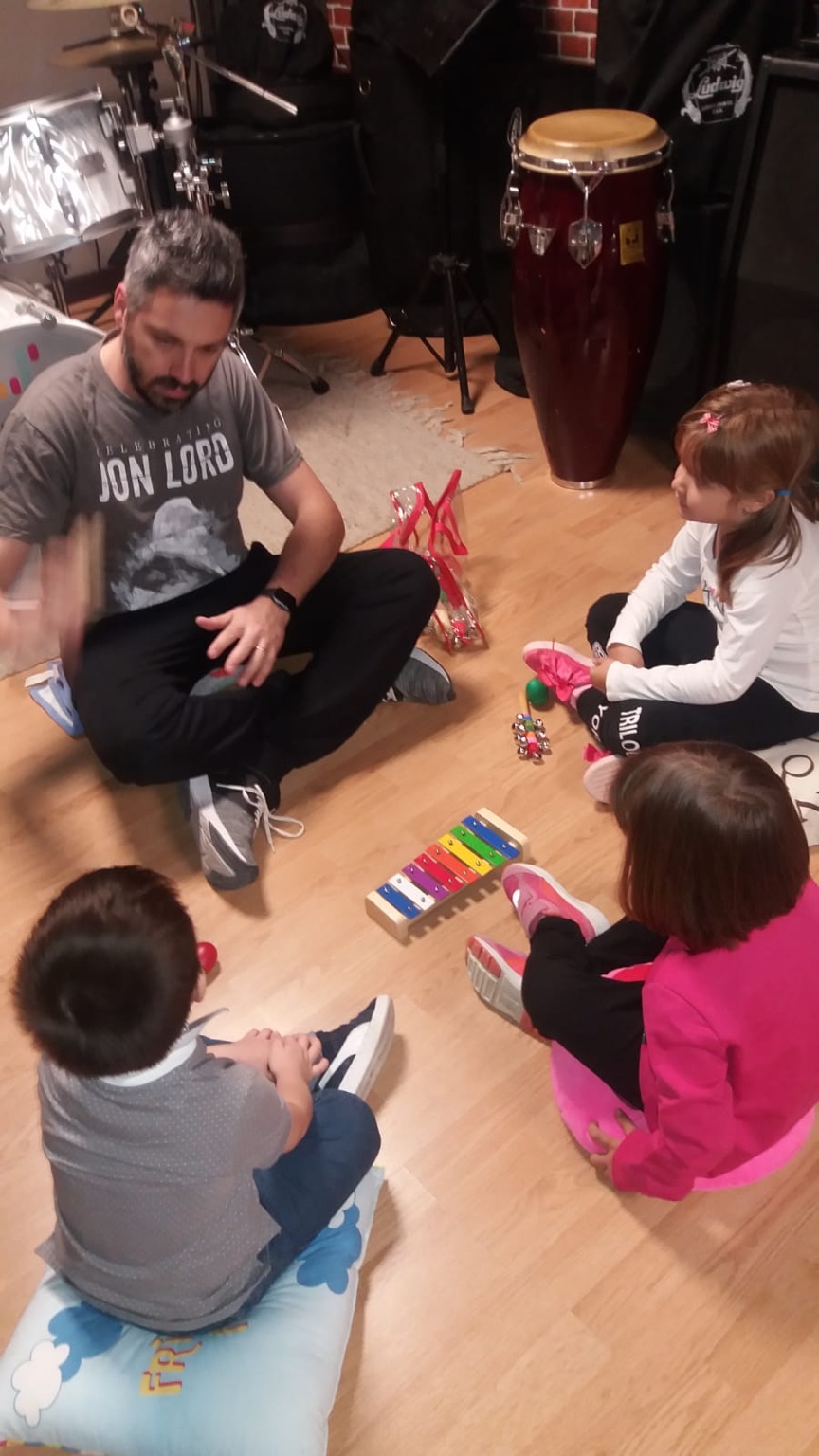 Armando Cacciato seduto in cerchio con i bambini durante un’attività musicale, tra strumentario Orff e percussioni